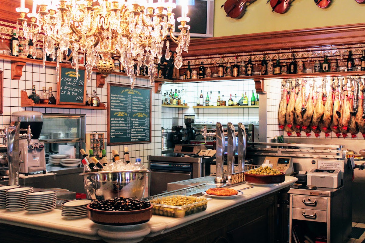 Interior de un bar típico español con grandes ollas de aceitunas en la parte superior de la barra y una gran araña de cristal en el techo.