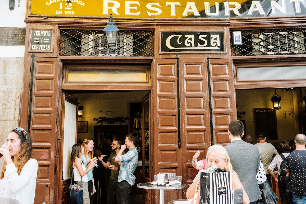 Exterior de un edificio de restaurante marrón con gente disfrutando de comida y bebida en una terraza al aire libre.