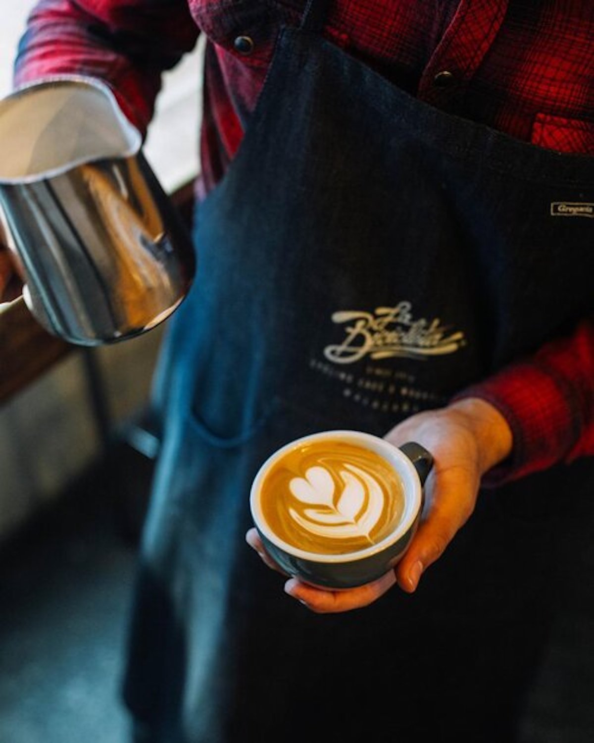 Barista sosteniendo una jarra plateada de leche y un café con leche