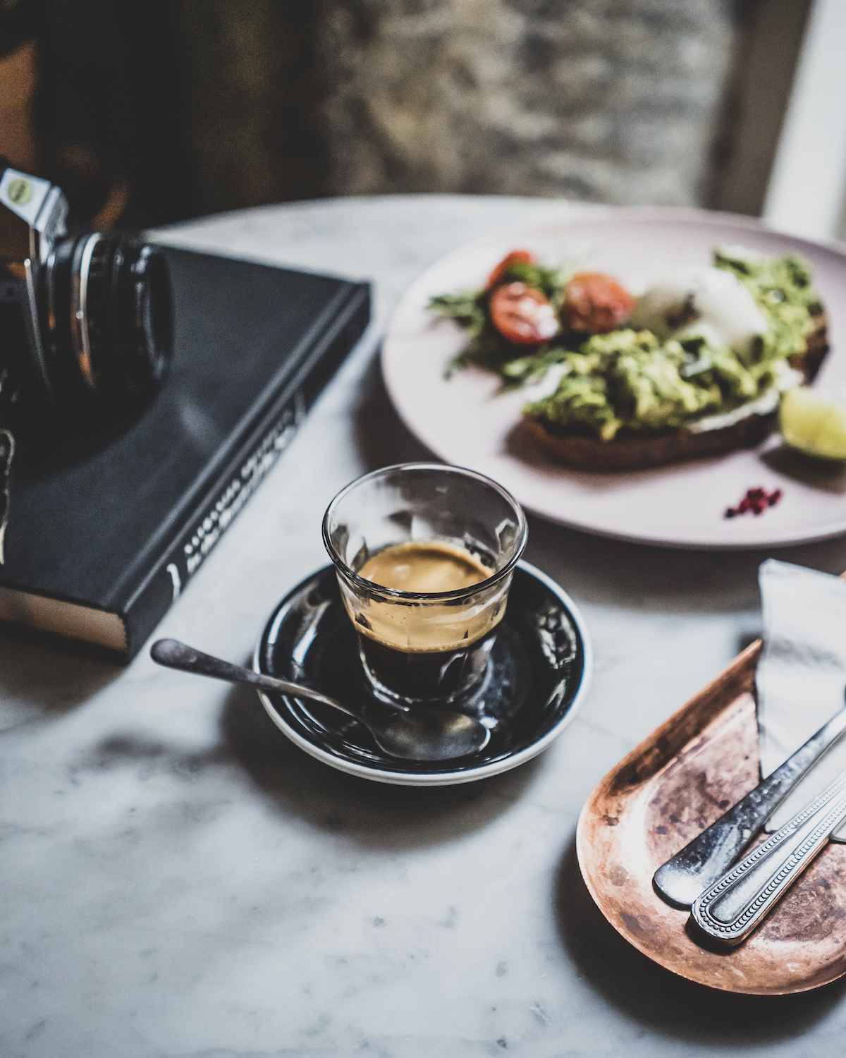 Vaso transparente de espresso junto a un libro y un plato de tostadas de aguacate sobre una mesa blanca.
