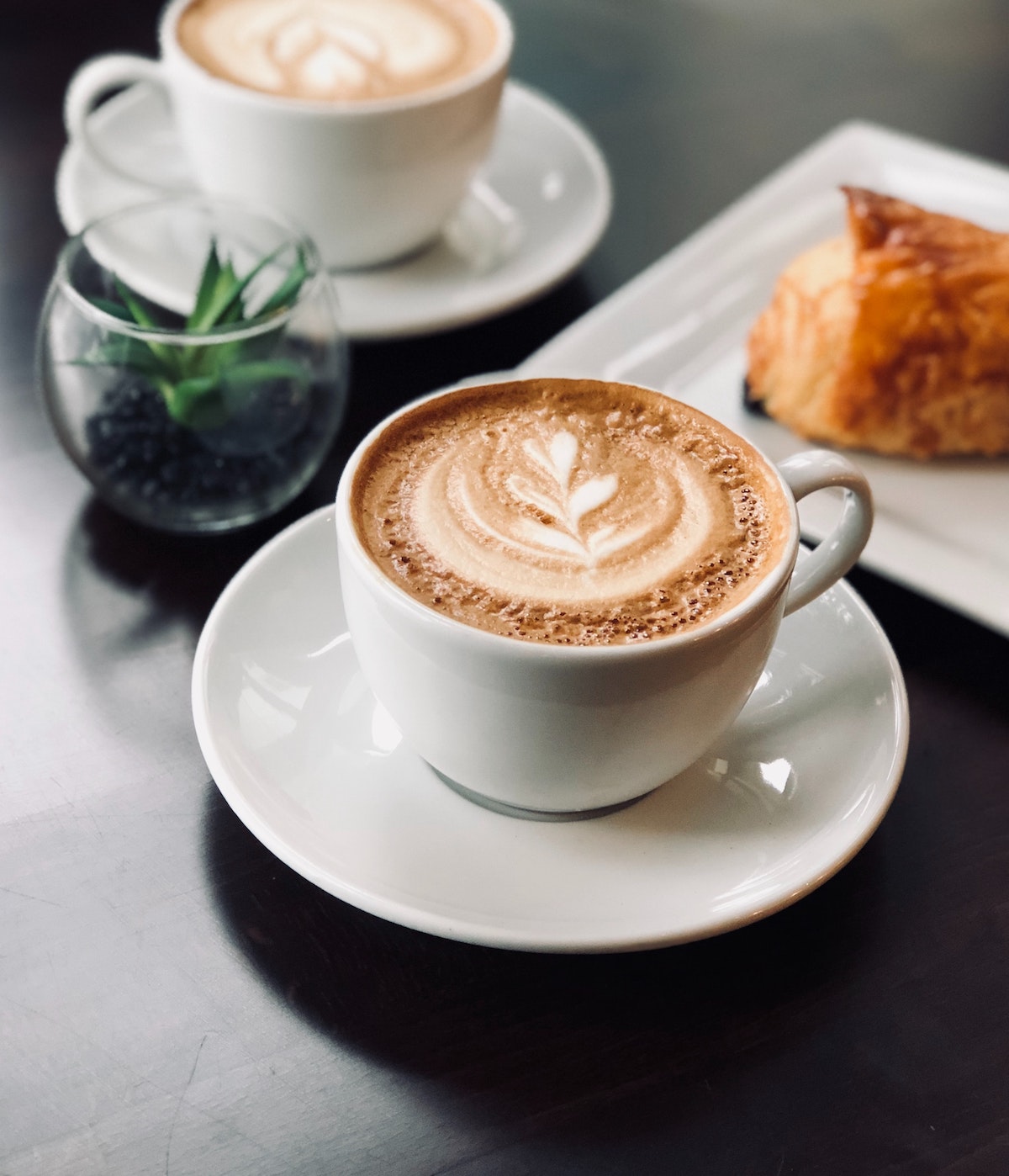 Dos cafés con leche en tazas blancas sobre una mesa junto a una pequeña maceta y un pastel en un plato blanco.