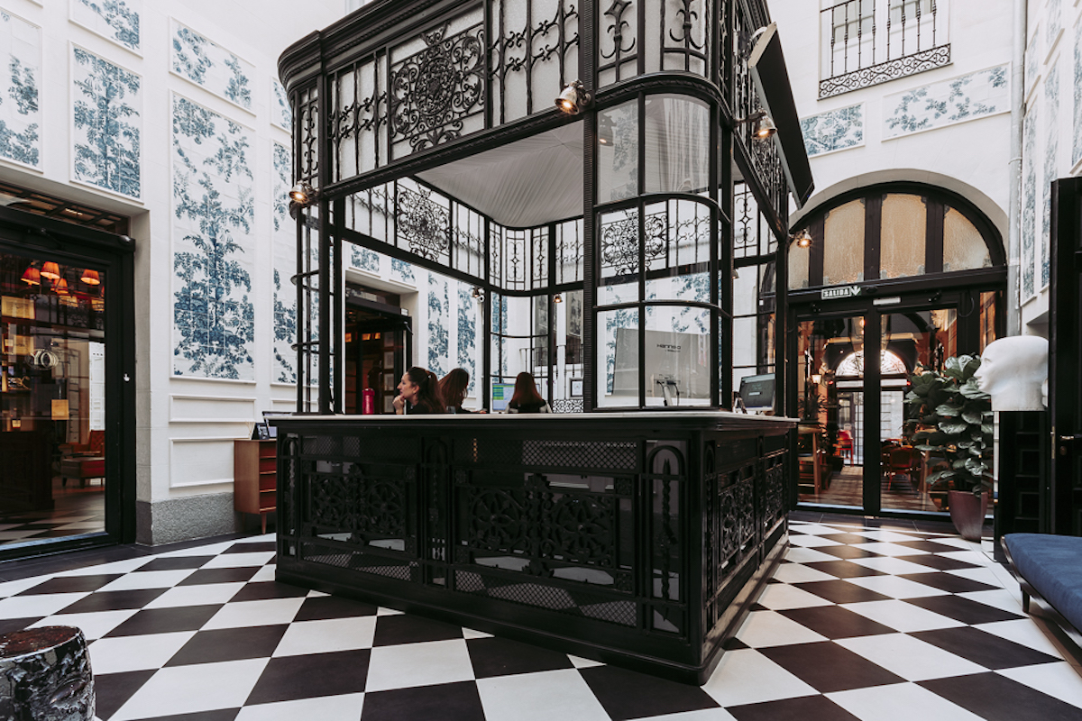 Vestíbulo del hotel con suelo a cuadros en blanco y negro y una recepción de hierro forjado ornamentada.