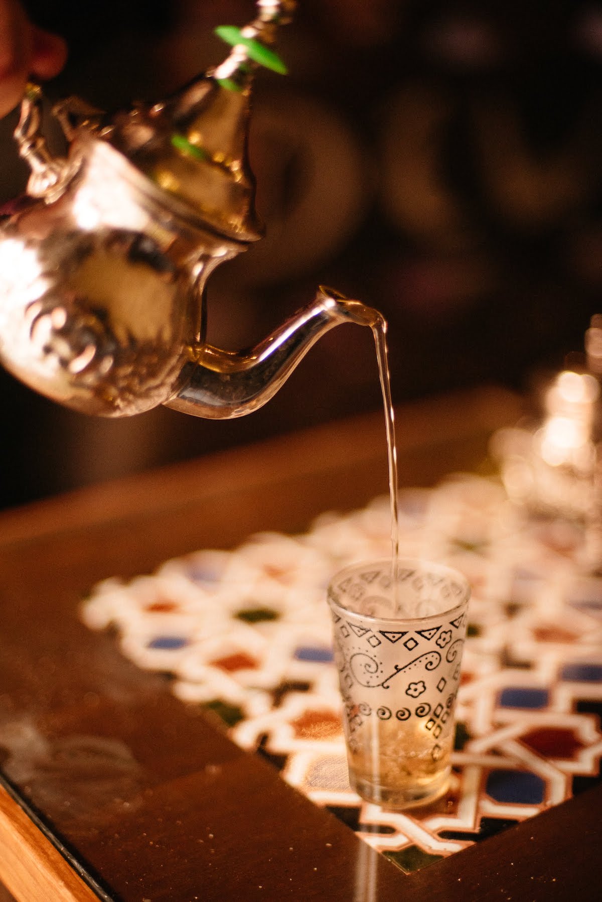 Té marroquí vertido de una tetera de metal a una taza de cristal sin asa sobre una mesa de azulejos.
