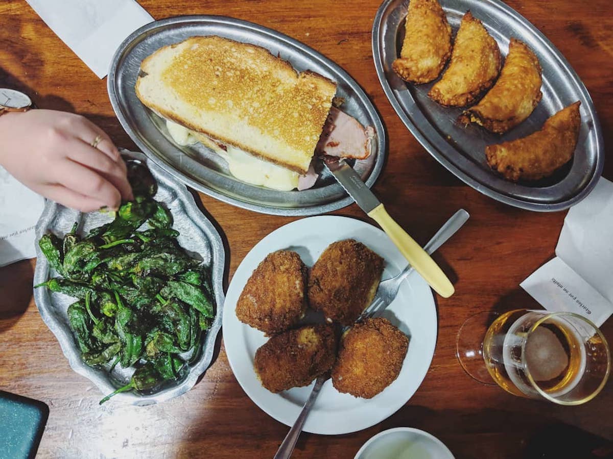 Toma aérea de un plato de pimientos verdes fritos, un bocadillo grande, croquetas fritas, una bandejita de mini empanadas y un vaso de cerveza.