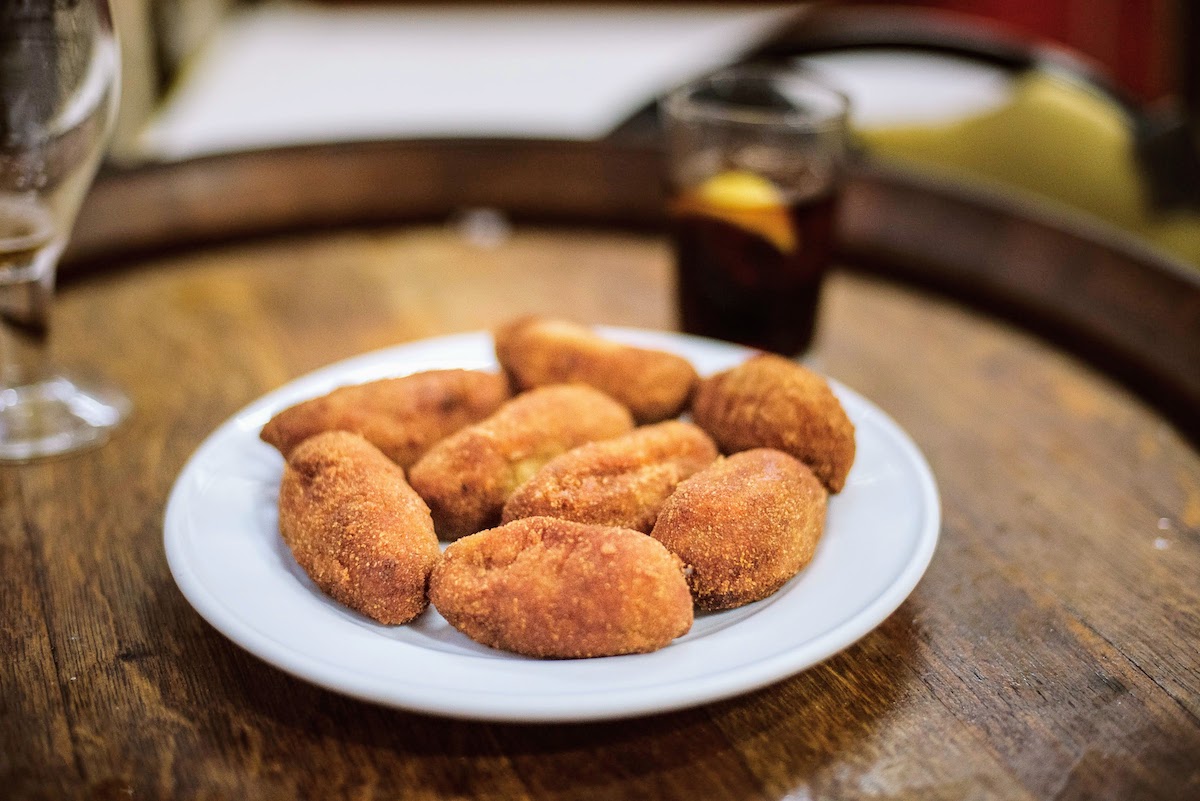 Varias croquetas en un plato blanco, con un vaso de vermut visible al fondo.