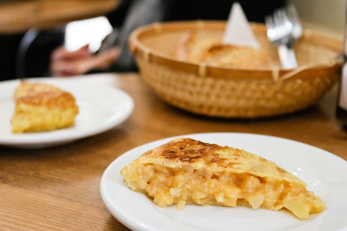 Dos rebanadas de tortilla de patata española en platos blancos separados, con una cesta de pan marrón al fondo.