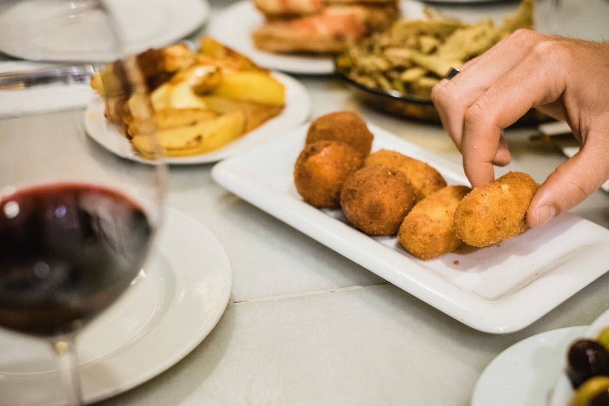 La mano de una persona cogiendo una croqueta frita de un pequeño plato blanco, con un vaso de vino tinto visible en primer plano.