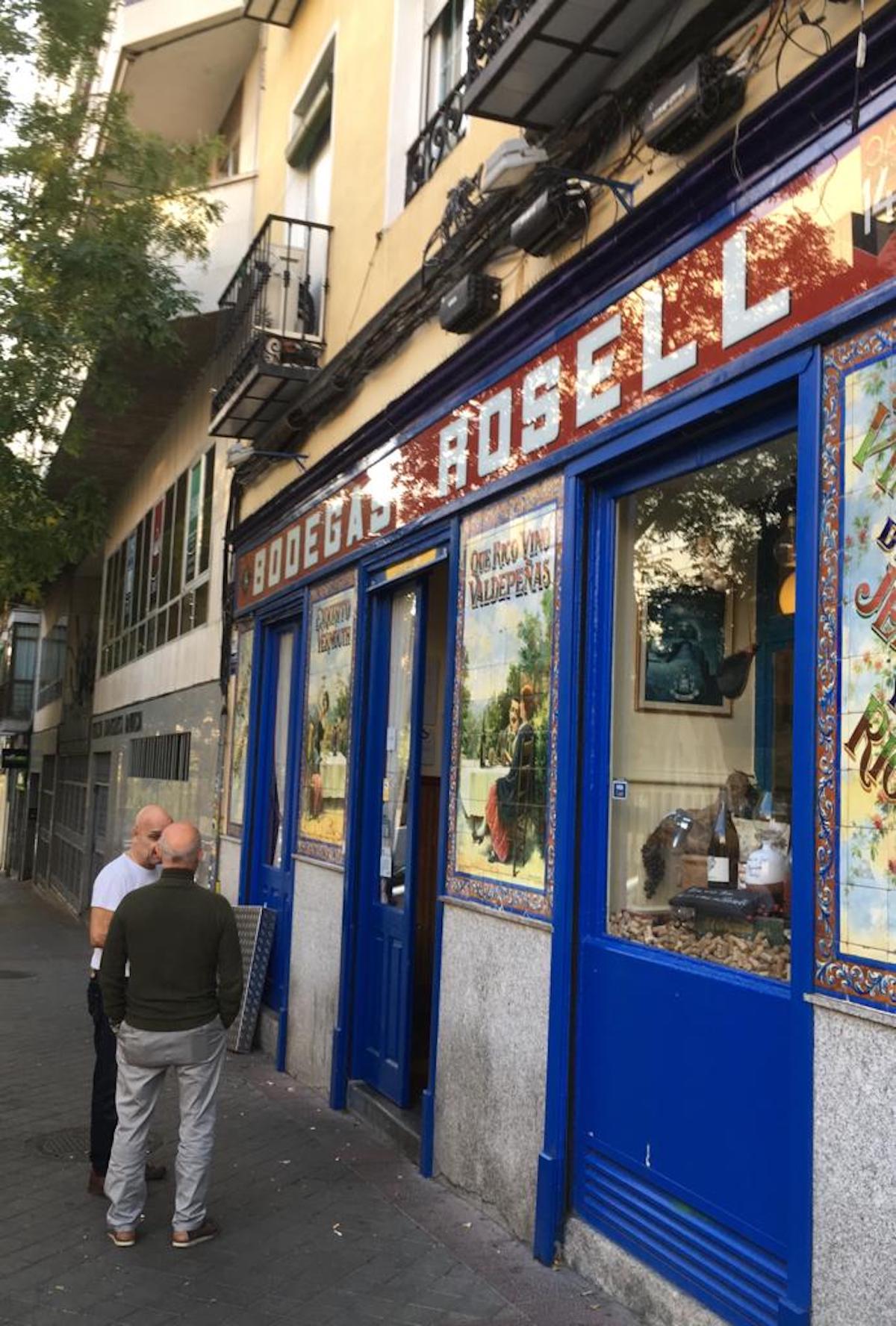 Dos hombres hablan fuera de un bar con paneles azules en las ventanas y anuncios de azulejos antiguos.