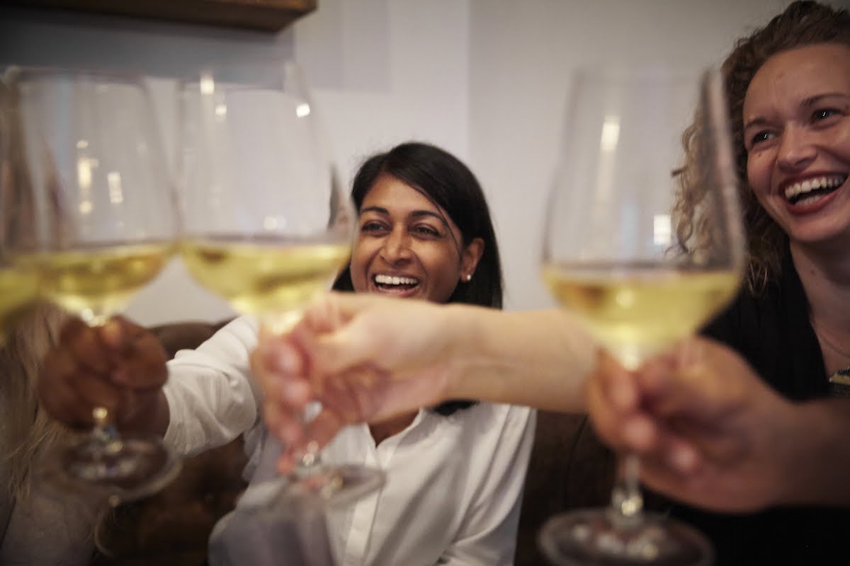Dos mujeres sonriendo mientras brindan con copas de vino blanco.
