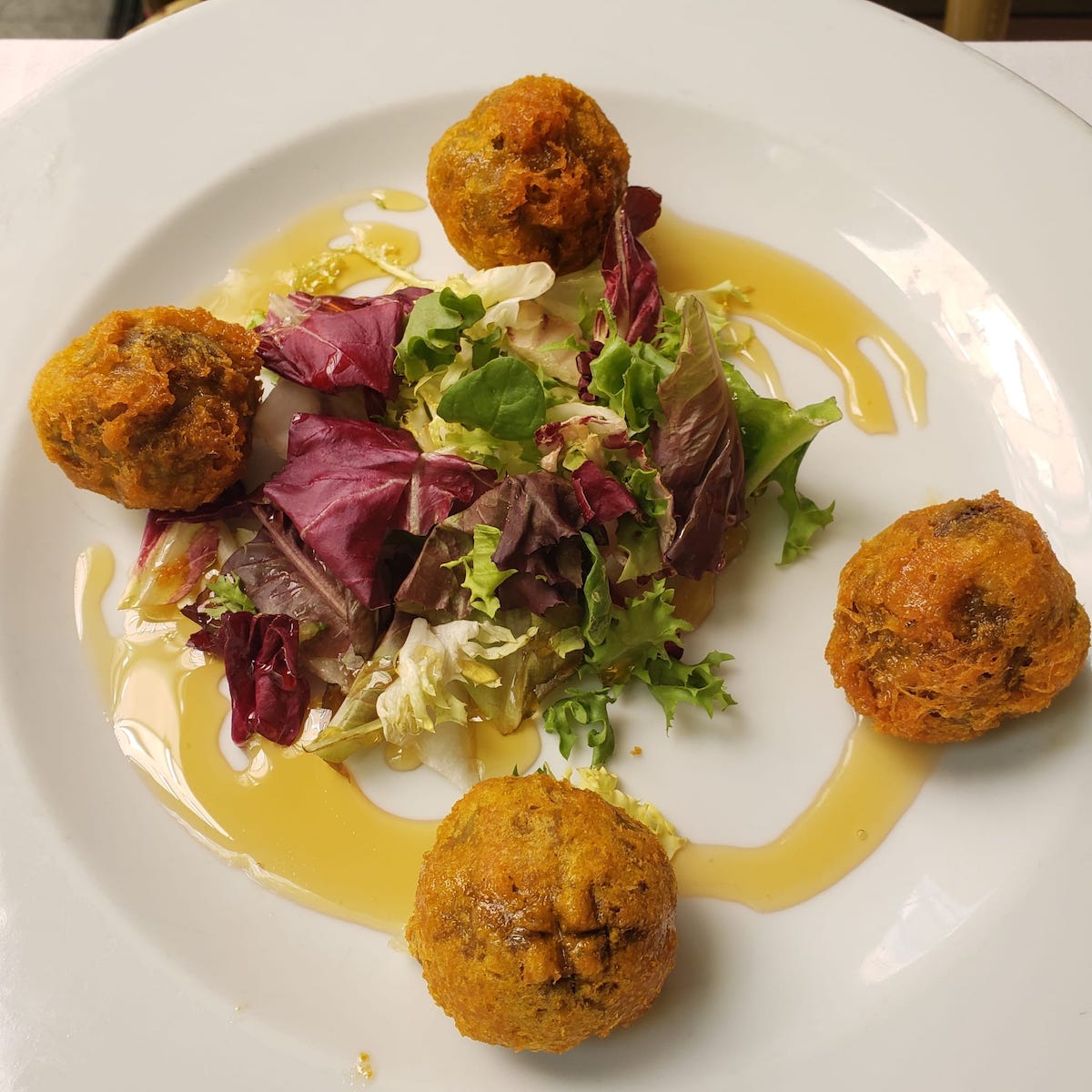 Cuatro croquetas fritas y una ensalada de hojas verdes en un plato blanco rociado con miel