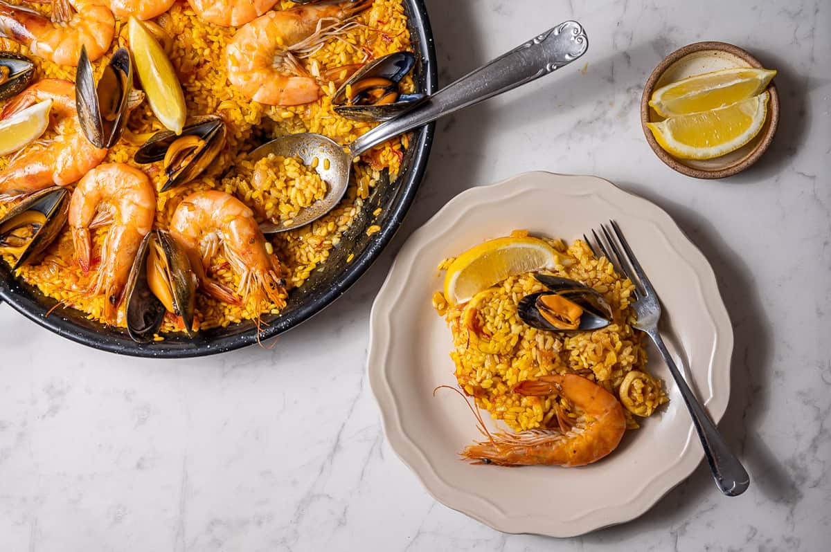 Toma aérea de una paella de marisco con una pequeña cantidad servida en un plato blanco.